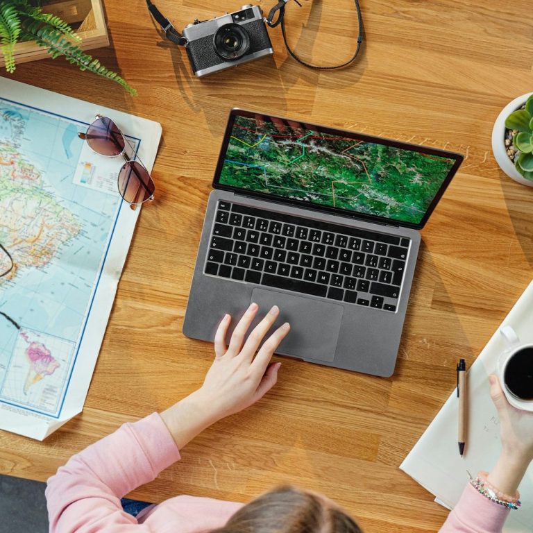 A woman putting together a plan for a customer A person using a laptop on a wooden table with a map, camera, and sunglasses nearby.