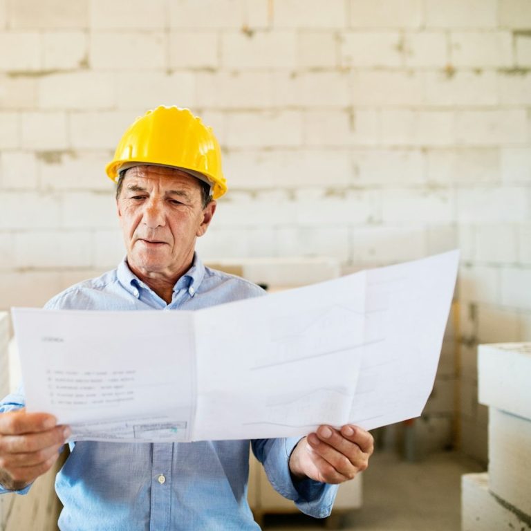 A man planning to upgrade a house An older man in a yellow hard hat reviews blueprints at a construction site.